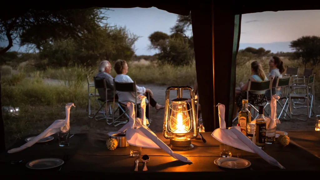 A cozy dinner setup inside a mobile camp in the Central Kalahari, Botswana, with a lantern illuminating the table, while guests enjoy a peaceful evening view outside.