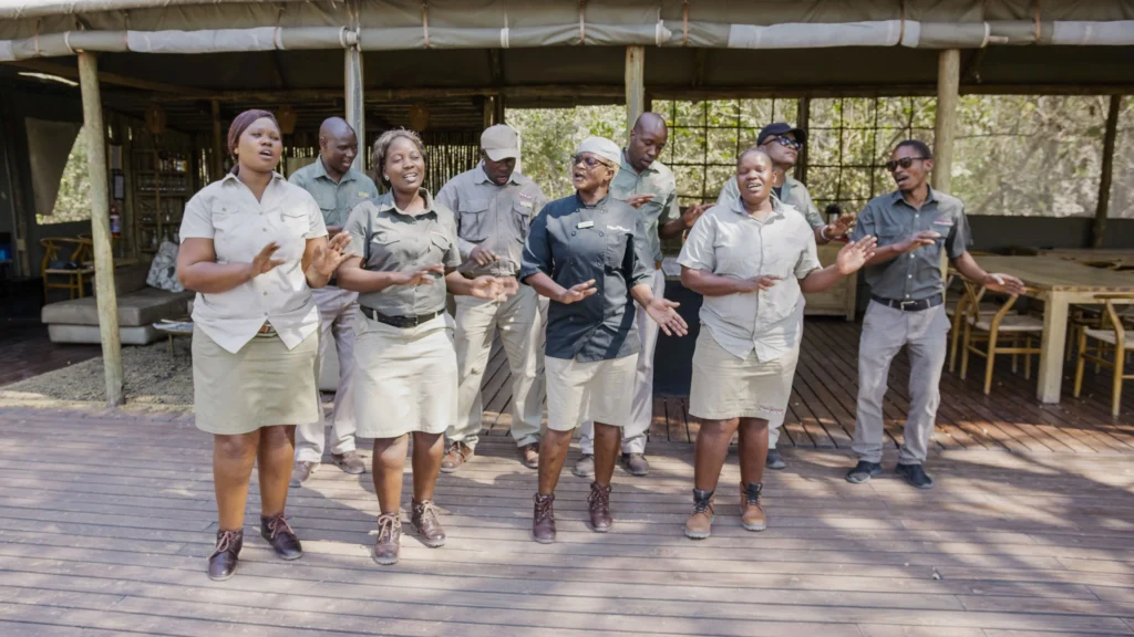 Warm and welcoming staff of O Bona Moremi Safari Lodge singing and clapping together on the wooden deck, sharing joyful energy with guests in the heart of the bush.