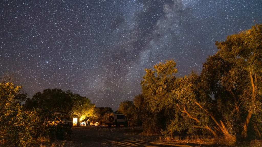 Mobile safari camp beneath the starry sky in Savuti, Botswana. The Milky Way stretches across the night sky, with camp lights illuminating the campsite.