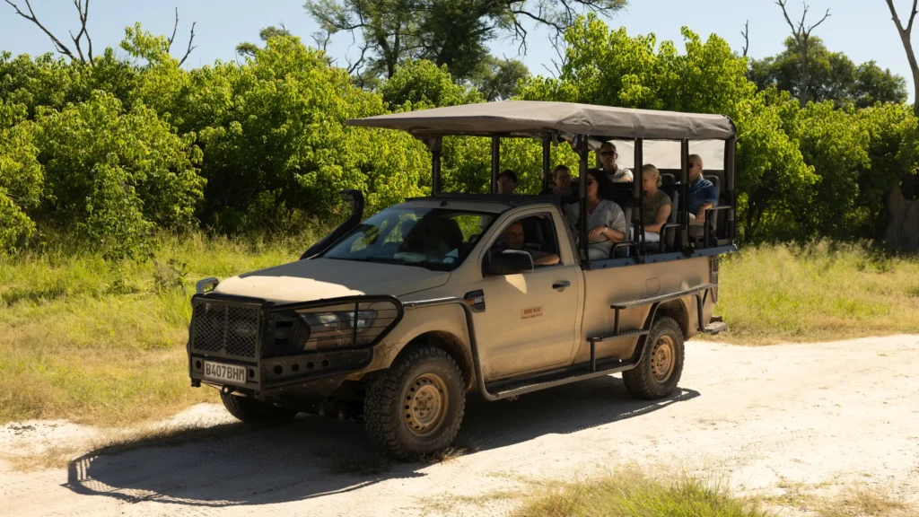 A group of travelers enjoys a game drive in the Khwai region of the Okavango Delta, observing wildlife from an open-air safari vehicle on a sunlit day.