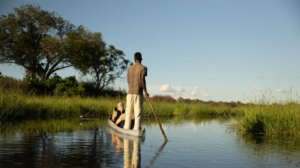 A mokoro guide poling through the peaceful waters of Khwai, Okavango Delta, while a guest captures the moment on their phone.