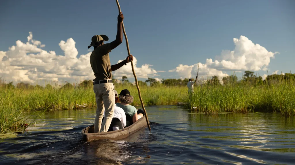 A mokoro guide steers a traditional canoe in the Khwai River, with passengers enjoying the serene waterway and surrounding lush greenery in Botswana's Okavango Delta.
