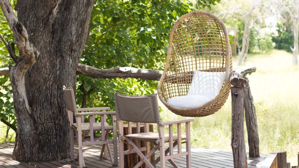 Peaceful deck at O Bona Moremi Safari Lodge with wooden chairs and a woven hanging chair shaded by a large tree â€“ a tranquil spot to relax and watch the bush come to life.