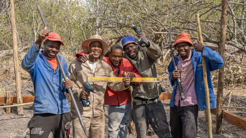 Construction staff of O Bona Moremi, smiling and posing for a photo with their tools, including levelers, drills, and picks, in a natural setting.